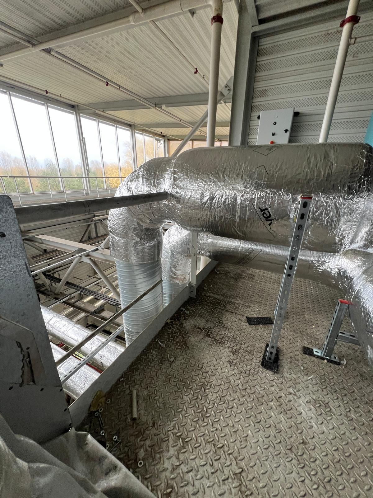 Des tuyaux isolés argentés reposent sur une plateforme dans un environnement industriel. Un escabeau se trouve à proximité.