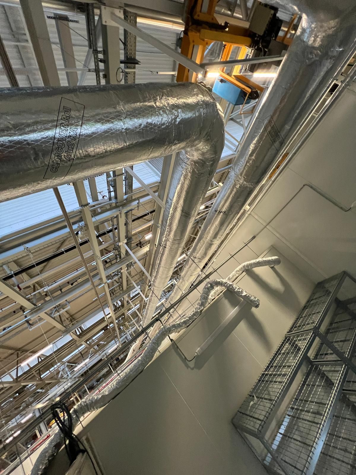Des tuyaux isolés argentés courent le long d'un haut plafond, avec des poutres métalliques et une vue imprenable sur le ciel.
