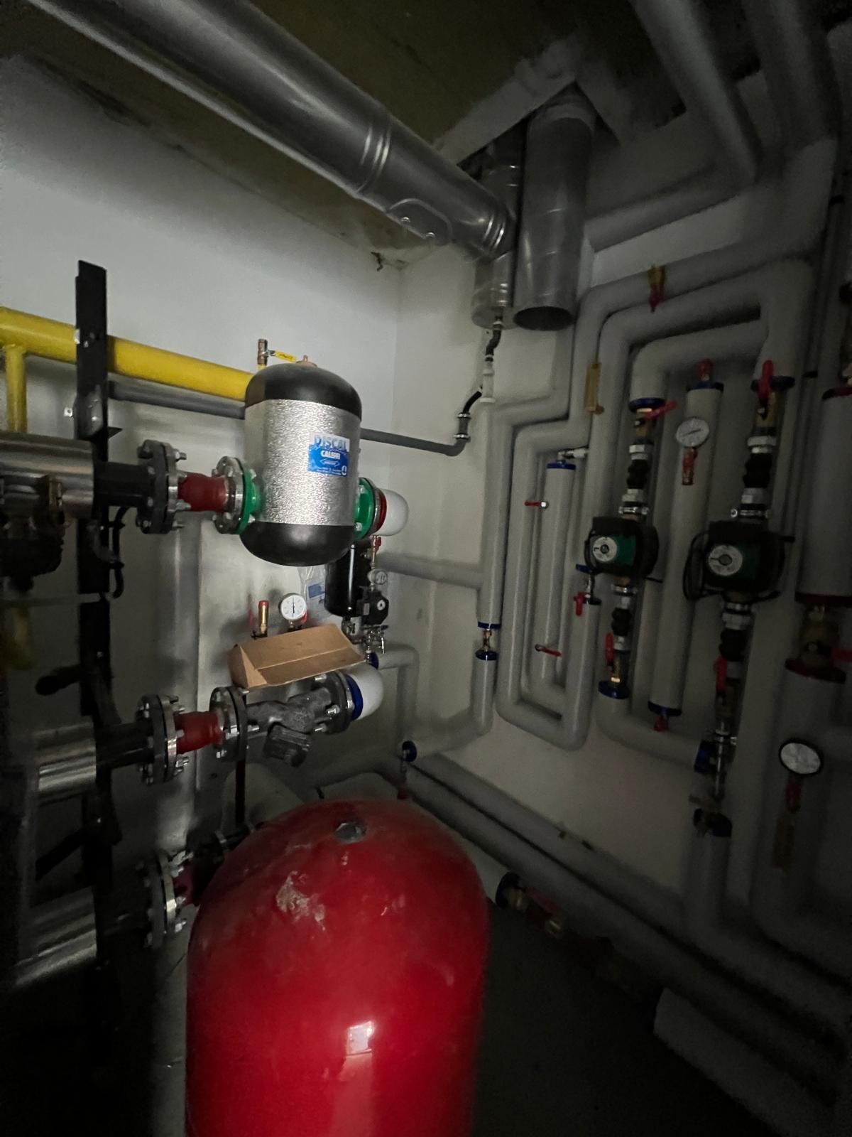 Local technique avec tuyauterie, réservoirs et pompes. On aperçoit des réservoirs rouges et noirs. Murs peints en blanc.