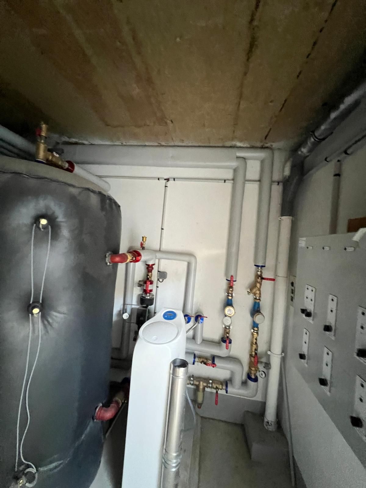 Système de chauffage dans un sous-sol : grand réservoir argenté, tuyaux blancs, manomètres et vannes contre des murs blancs.
