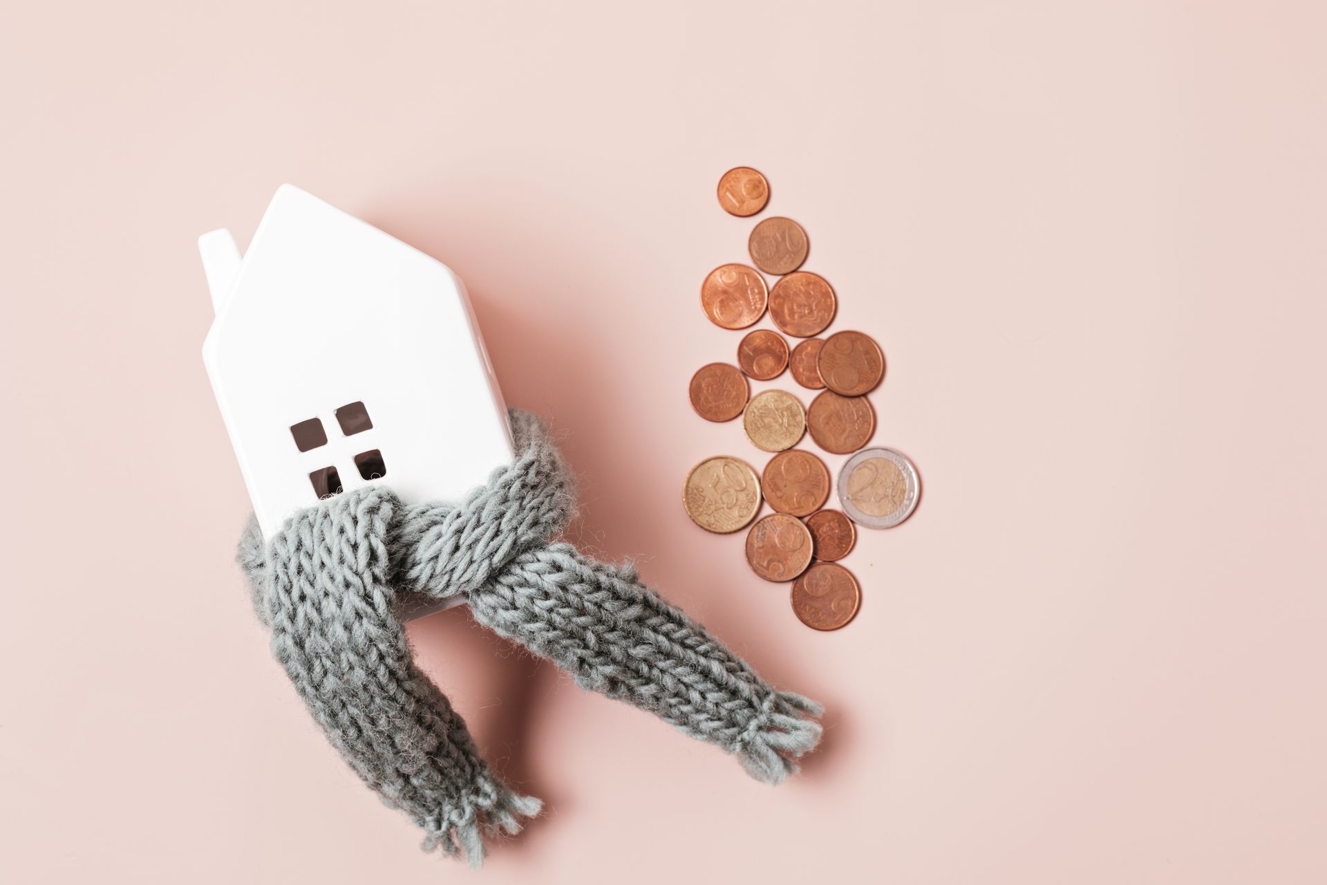 Maison miniature blanche portant une écharpe grise à côté d'un tas de pièces de cuivre sur fond rose.