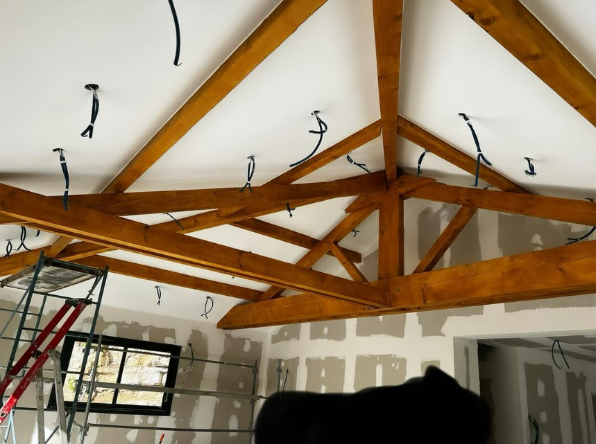 Vue intérieure d'un plafond de chantier avec poutres en bois, câbles électriques et plaques de plâtre.
