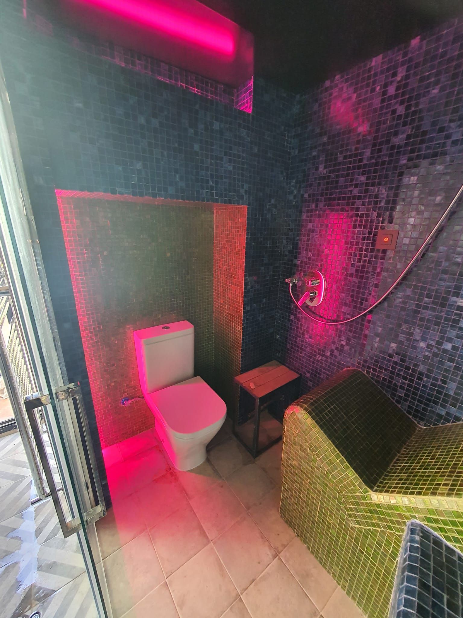 Un baño con paredes de mosaico, un inodoro, un pequeño taburete e iluminación rosa.