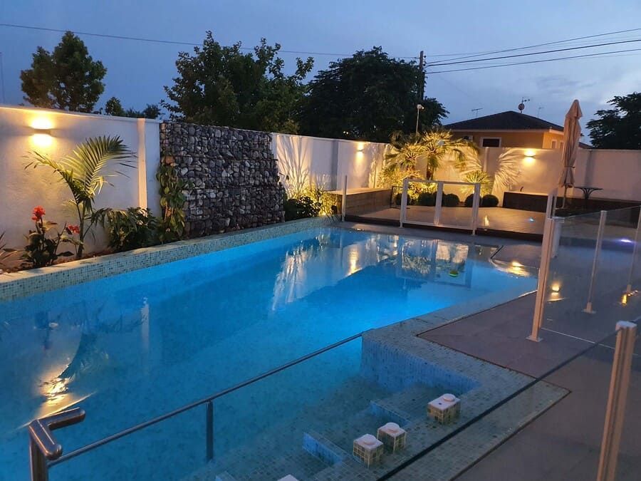 Vista nocturna de una piscina con iluminación azul. Cascada y jardín iluminado sobre una pared blanca.