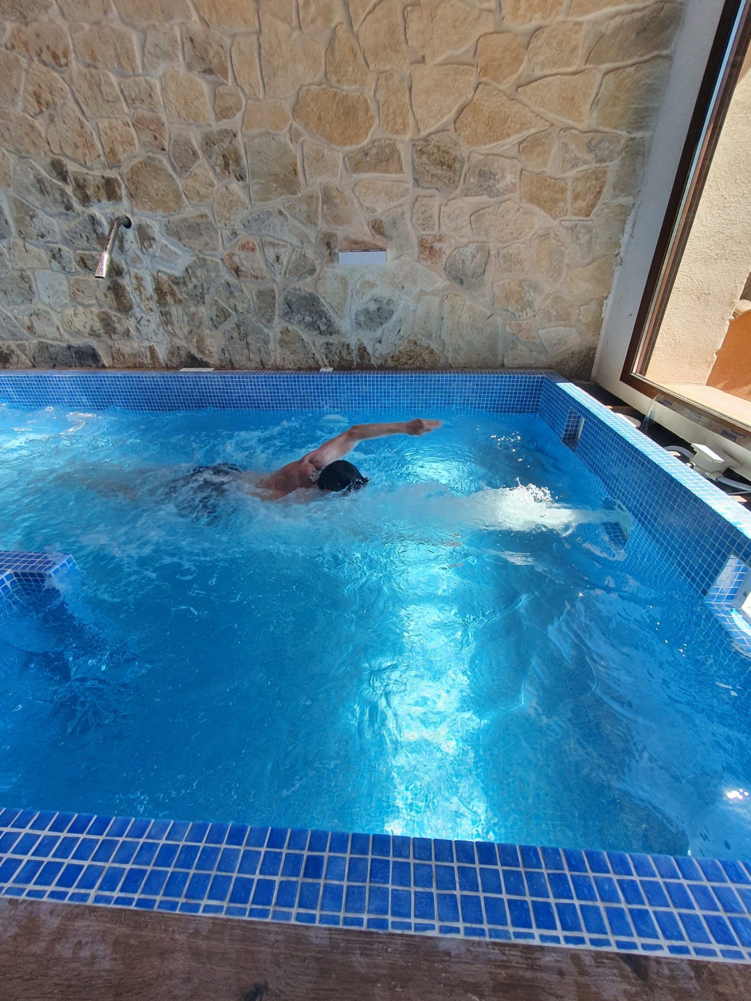 Una persona nadando en una piscina de azulejos azules bajo un muro de piedra.