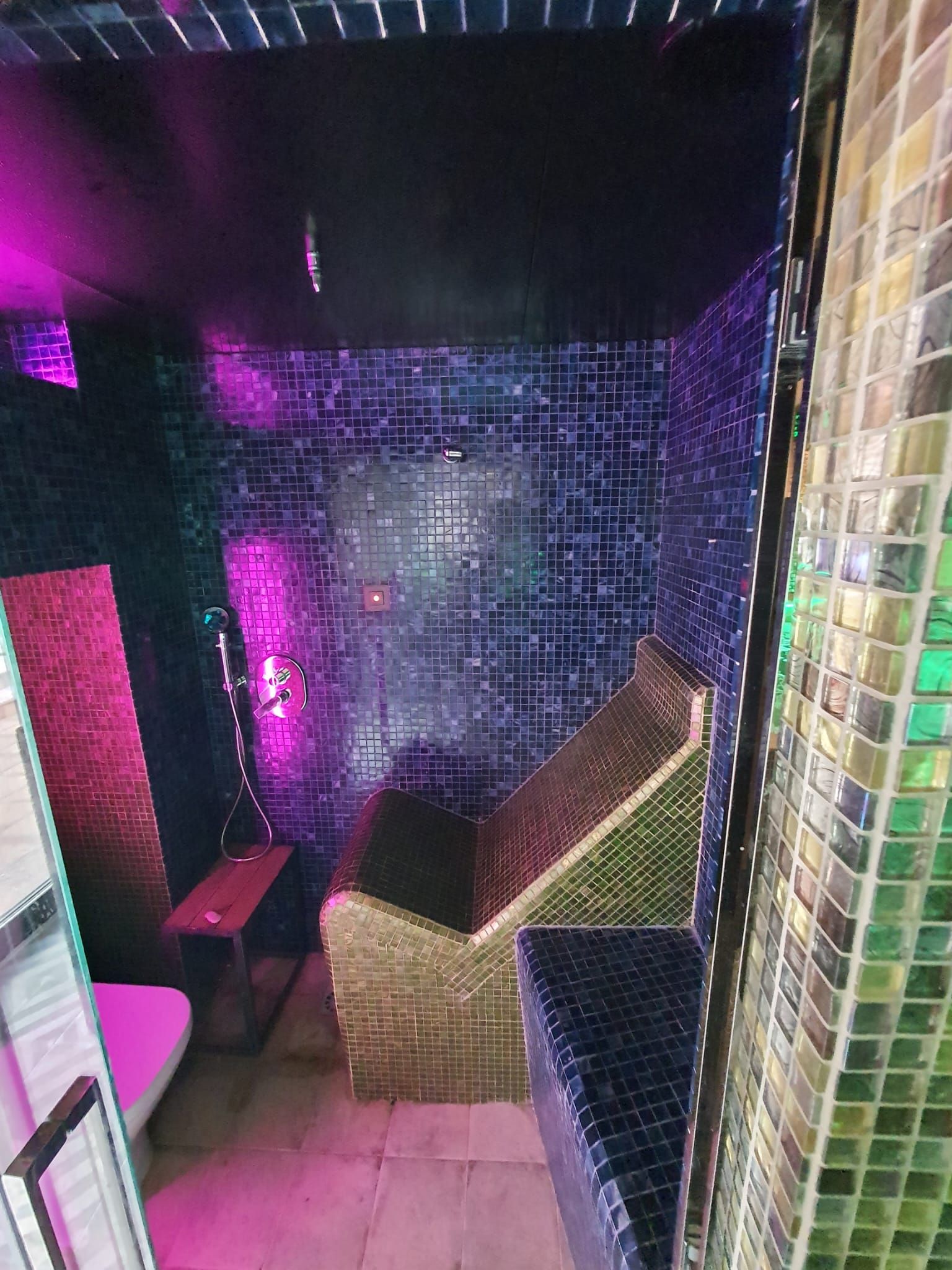 Una pequeña sala de temática disco con azulejos de espejo. Luces rosas y azules iluminan bancos y paredes.