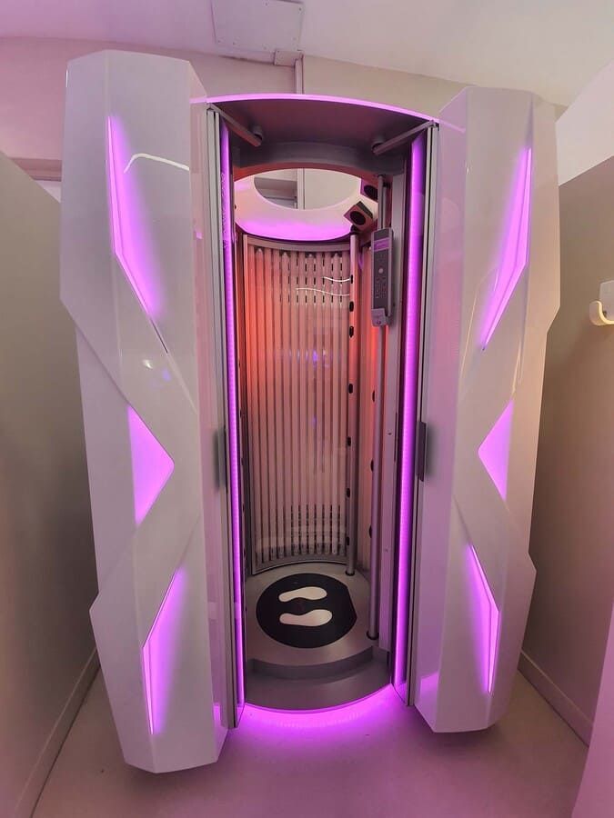 Cabina de bronceado blanca con luces rosas. Abierta, mostrando las bombillas interiores y la guía de pasos en el suelo.