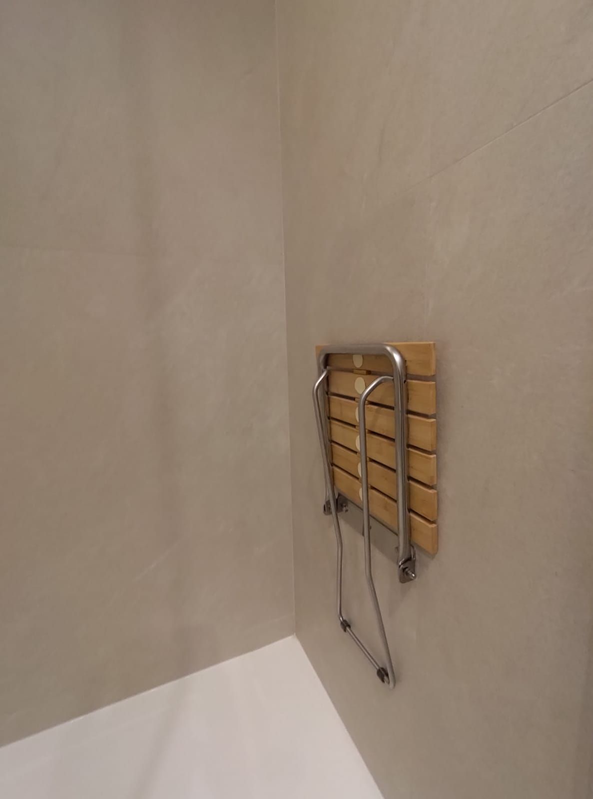 Asiento de ducha abatible de madera, montado en una pared beige en una esquina. Estructura de acero inoxidable con el asiento plegado.