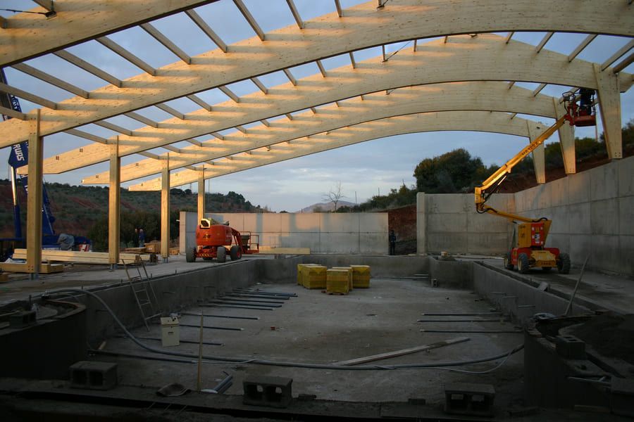 Obra de construcción de piscina bajo estructura de techo de madera; trabajadores en plataformas elevadoras.
