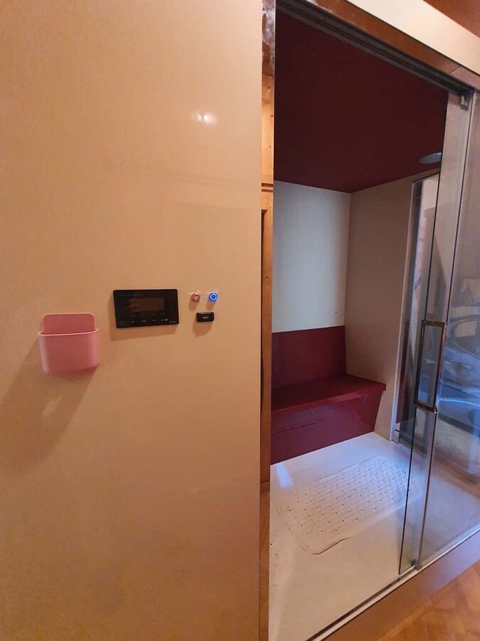 Una pequeña sauna con un banco y un techo de color rojo, al lado de una pared con un panel de control y un contenedor de almacenamiento de color rosa.