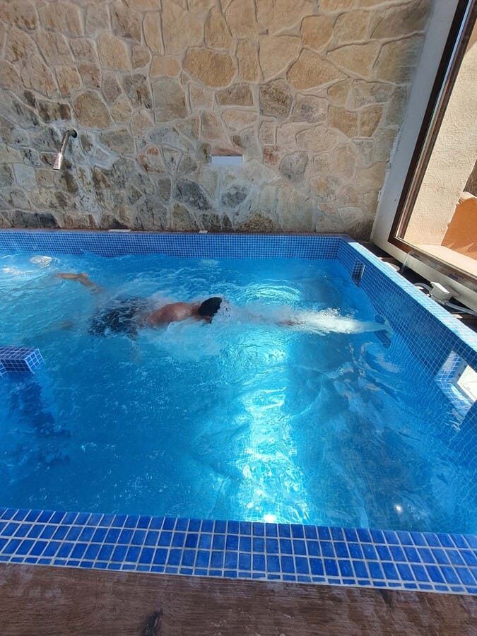 Persona nadando estilo libre en una pequeña piscina cubierta con azulejos azules y pared de piedra de fondo.