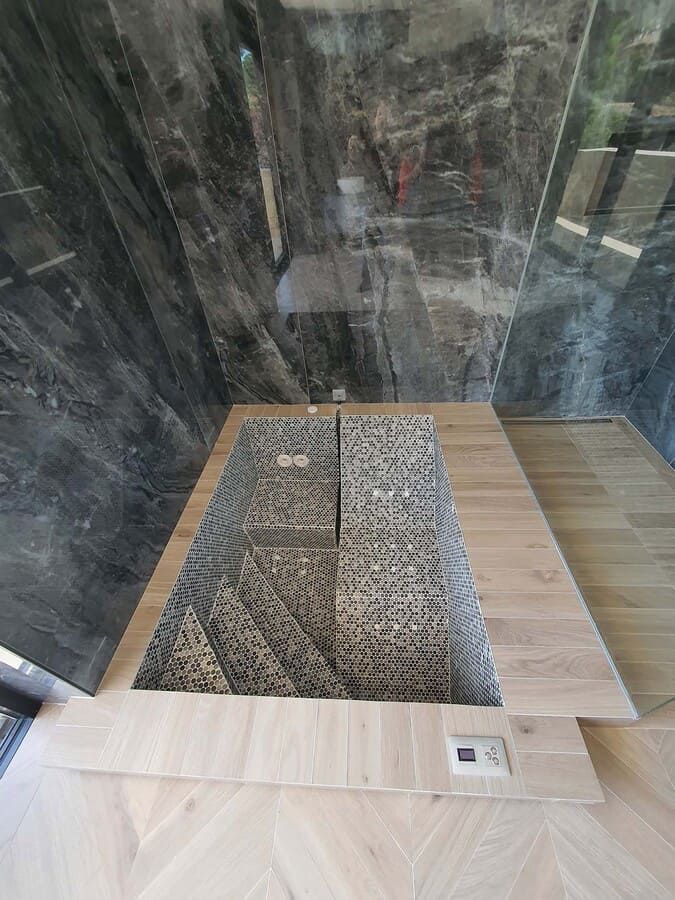 Baño de lujo con piscina de inmersión con azulejos de piedra, terraza de madera y pared de mármol oscuro.