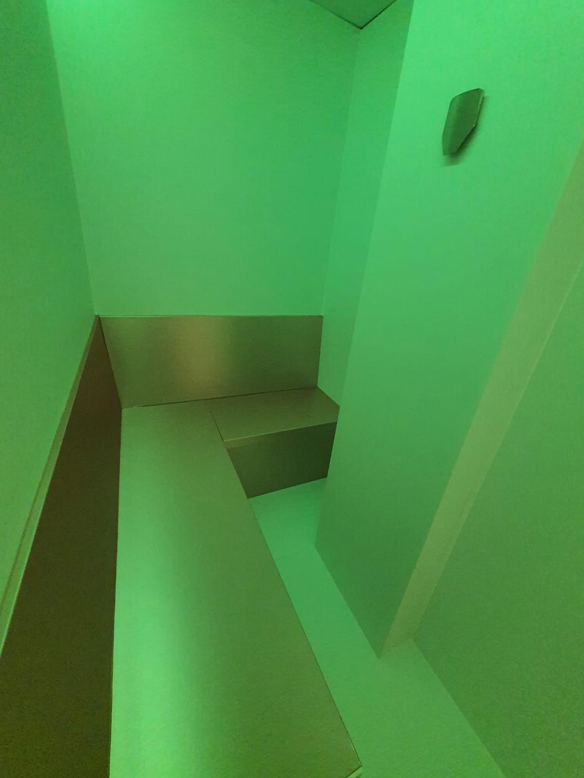 Habitación angular con luz verde y un banco de metal. Hay un pequeño objeto en la pared.