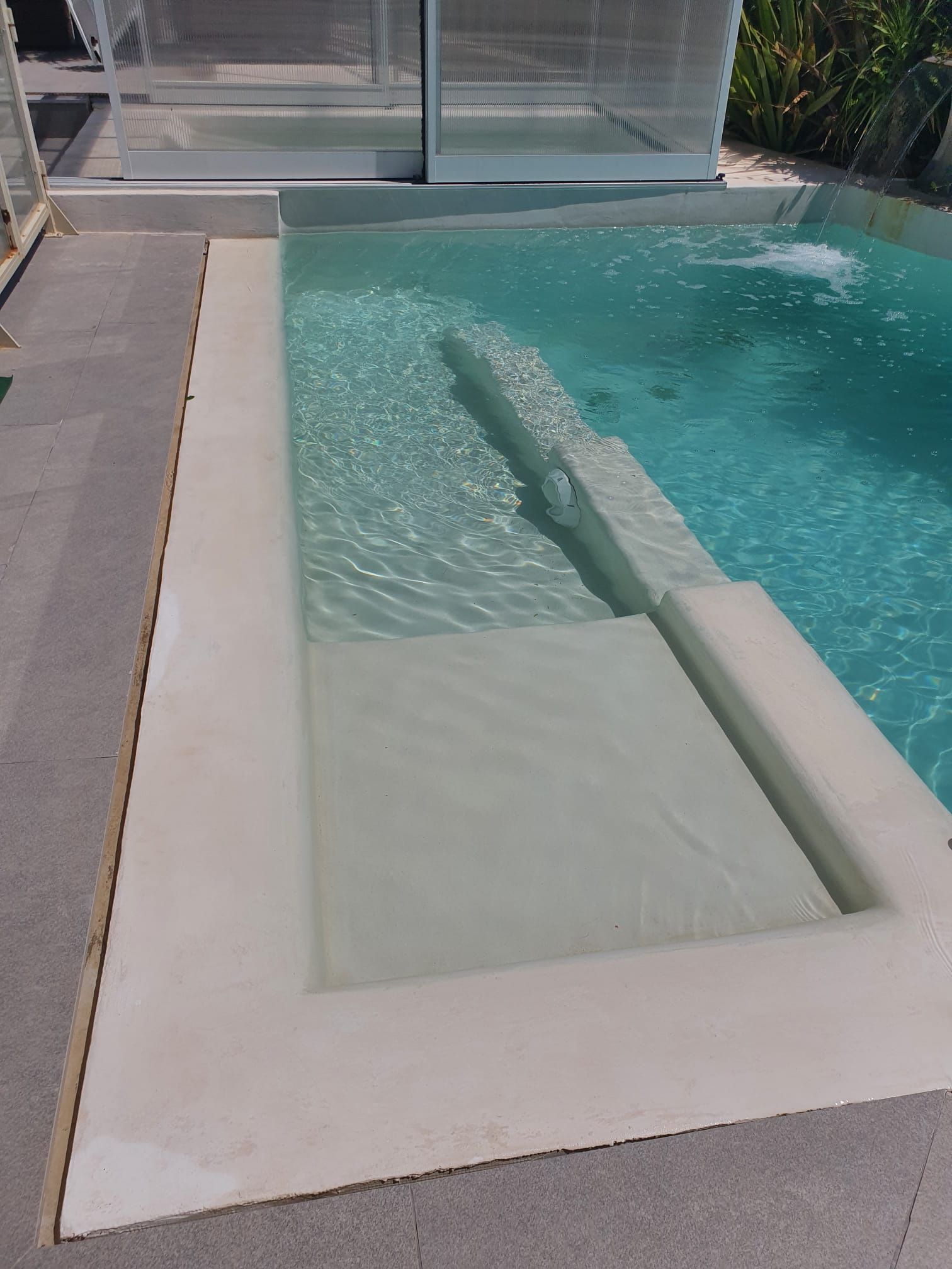 Una piscina moderna con juegos de agua y escalones integrados. El hormigón beige rodea el agua cristalina.