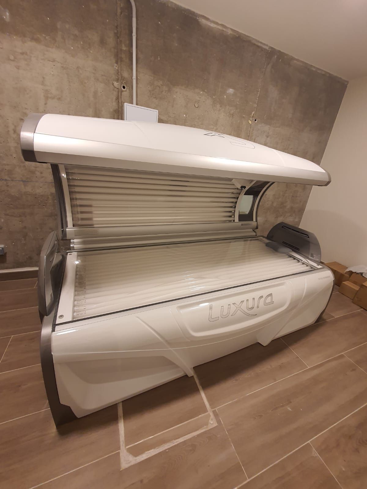 Cama de bronceado Luxura blanca en una habitación con una pared gris claro y un piso de baldosas marrones.