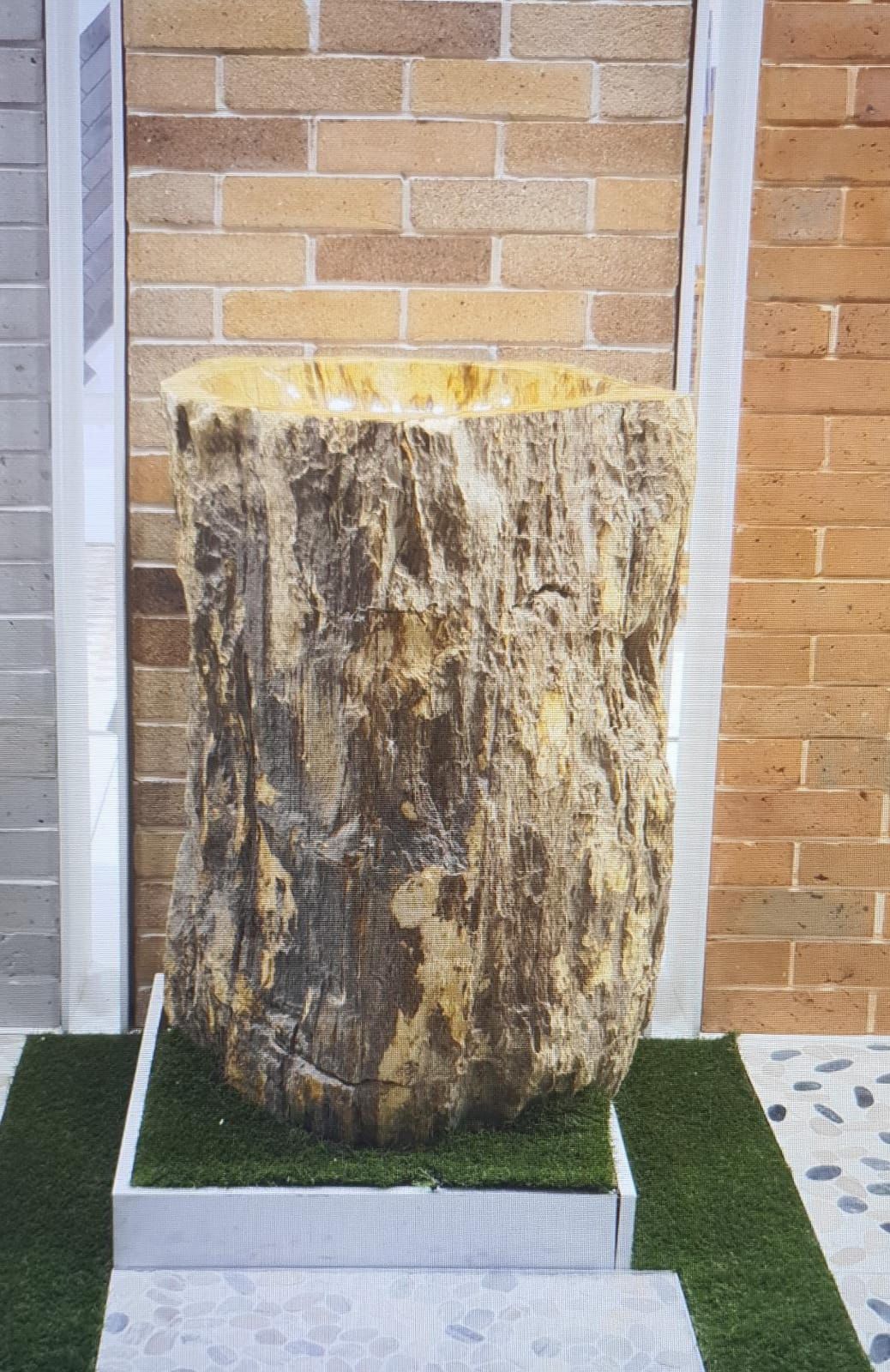 Una gran jardinera con forma de tocón de árbol, de color tostado y marrón, reposa sobre césped artificial verde contra paredes de ladrillo.