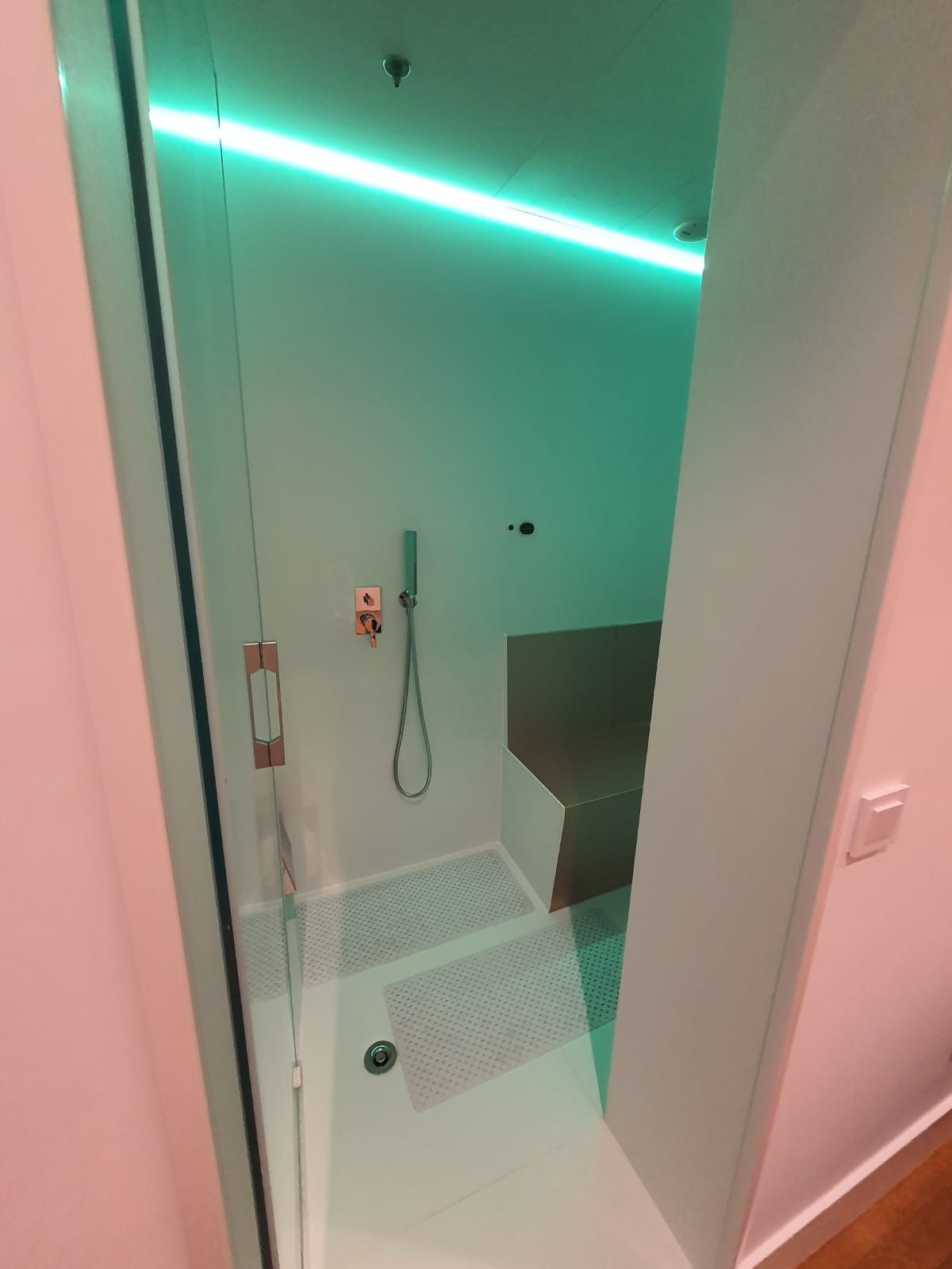 Cabina de ducha con puerta de cristal, iluminación cenital de color verde y banco incorporado.