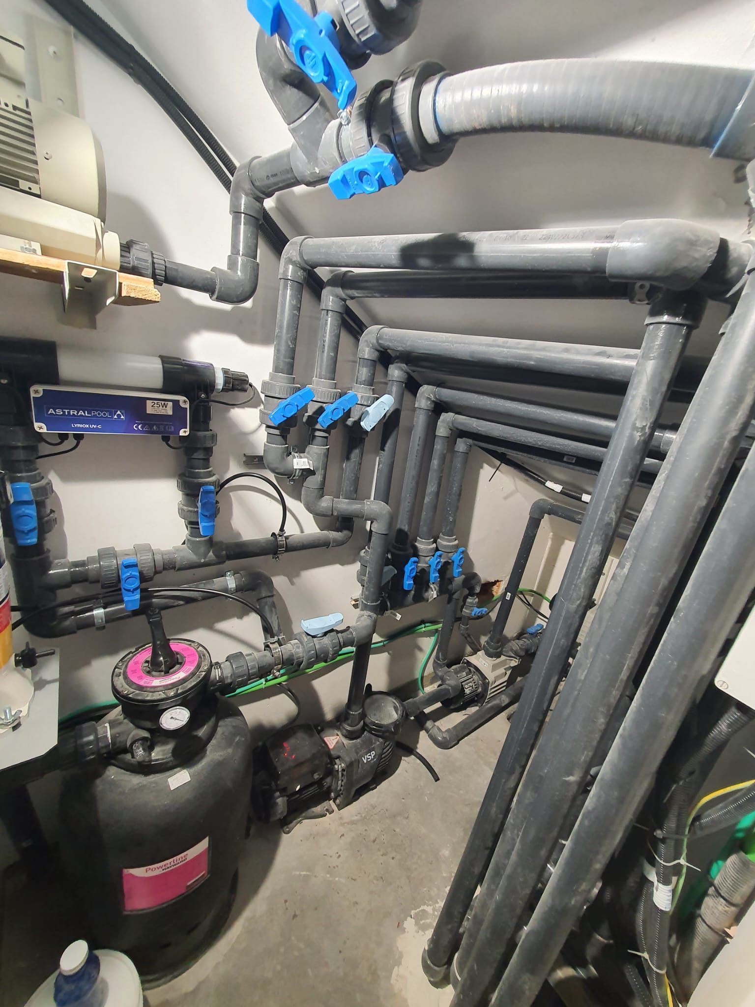 Sala de equipos de piscina con tuberías negras y azules, válvulas y filtro.
