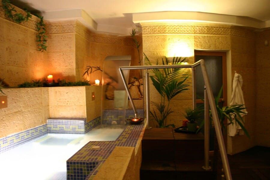 Interior de spa con bañera de hidromasaje de azulejos, velas, plantas y pasamanos.