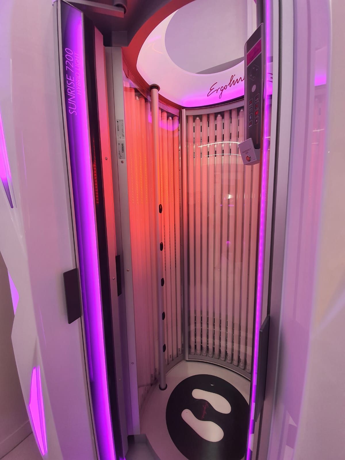 Cama de bronceado con tubos de luz verticales en su interior, luces de color violeta y naranja, exterior de color blanco.