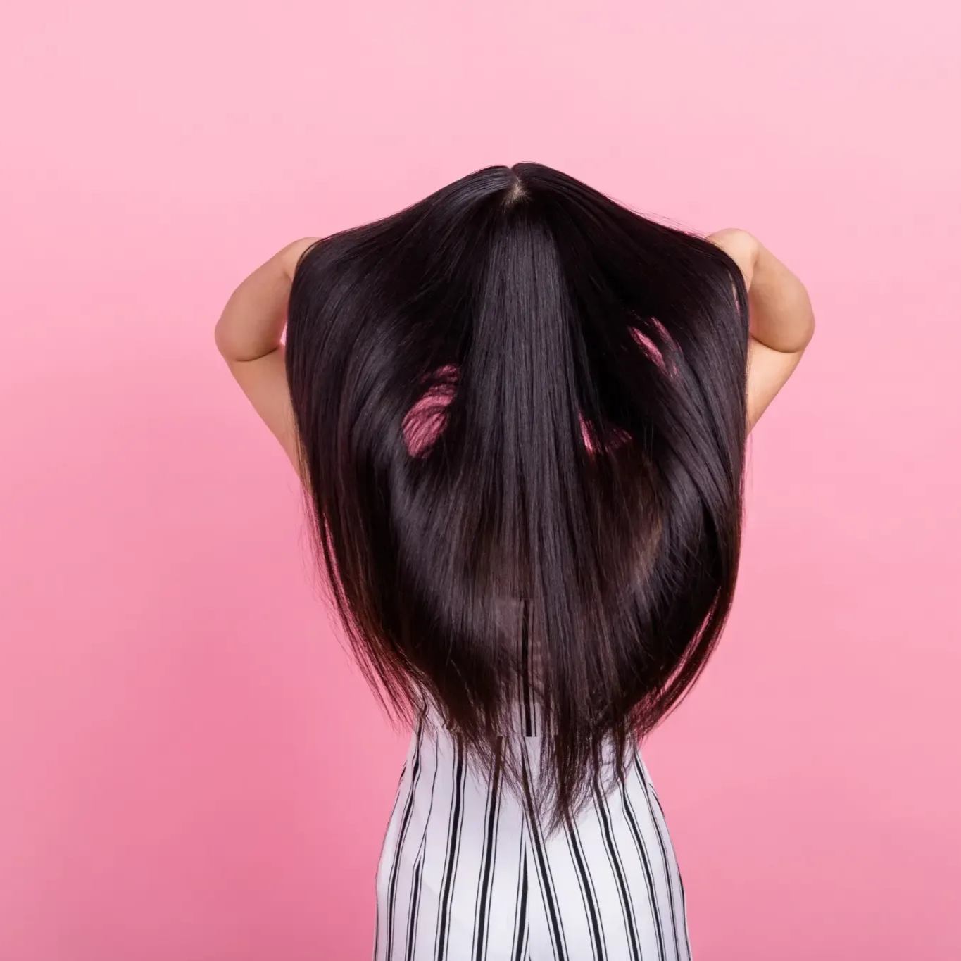 Una persona aparece de pie frente a un fondo rosa, sosteniendo con ambas manos una larga melena lisa y oscura.