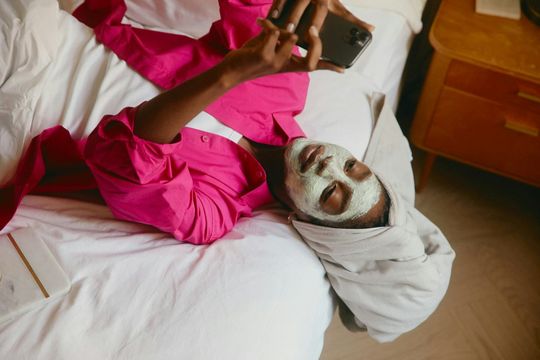 Person in a bright pink robe and towel taking a selfie while lying on a bed