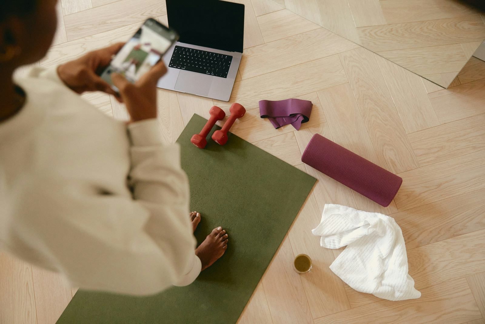 Person on a phone beside a laptop, yoga mat, dumbbells, water bottle, towel, and foam roller on the floor