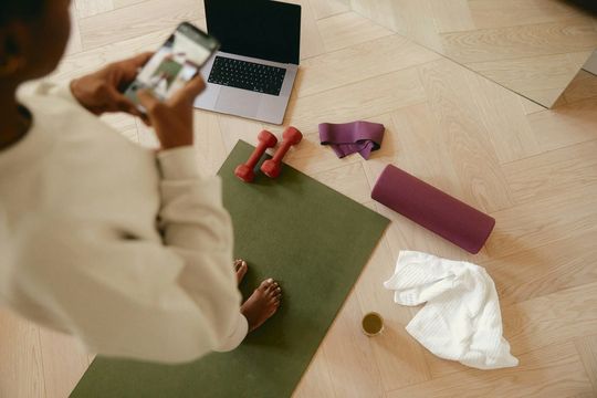 Person on a phone beside a laptop, yoga mat, dumbbells, water bottle, towel, and foam roller on the floor
