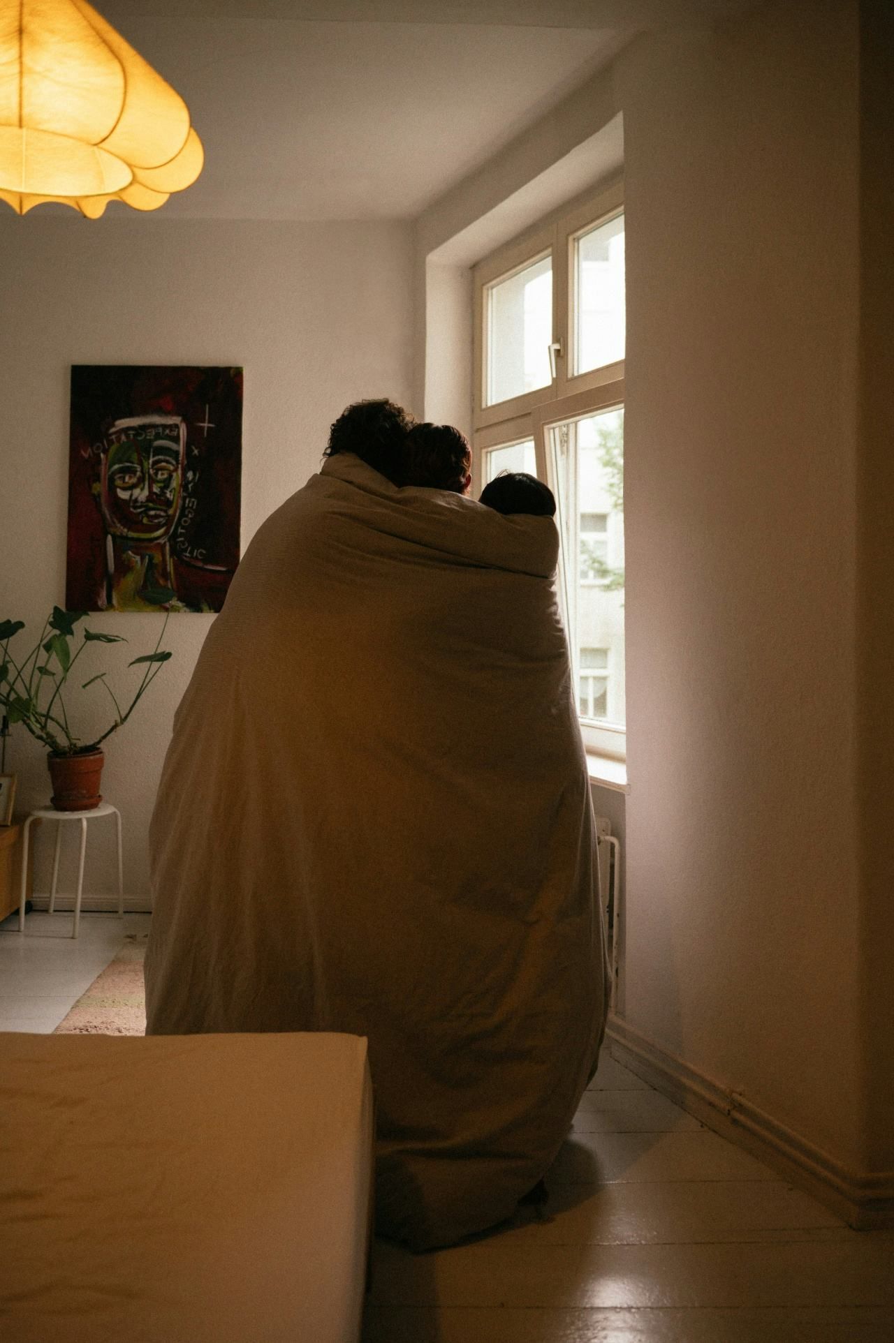 Two people wrapped in a tan blanket sit by a bright window in a cozy room.