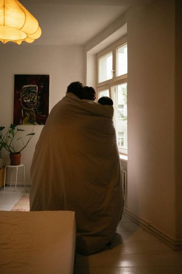 Two people wrapped in a tan blanket sit by a bright window in a cozy room.
