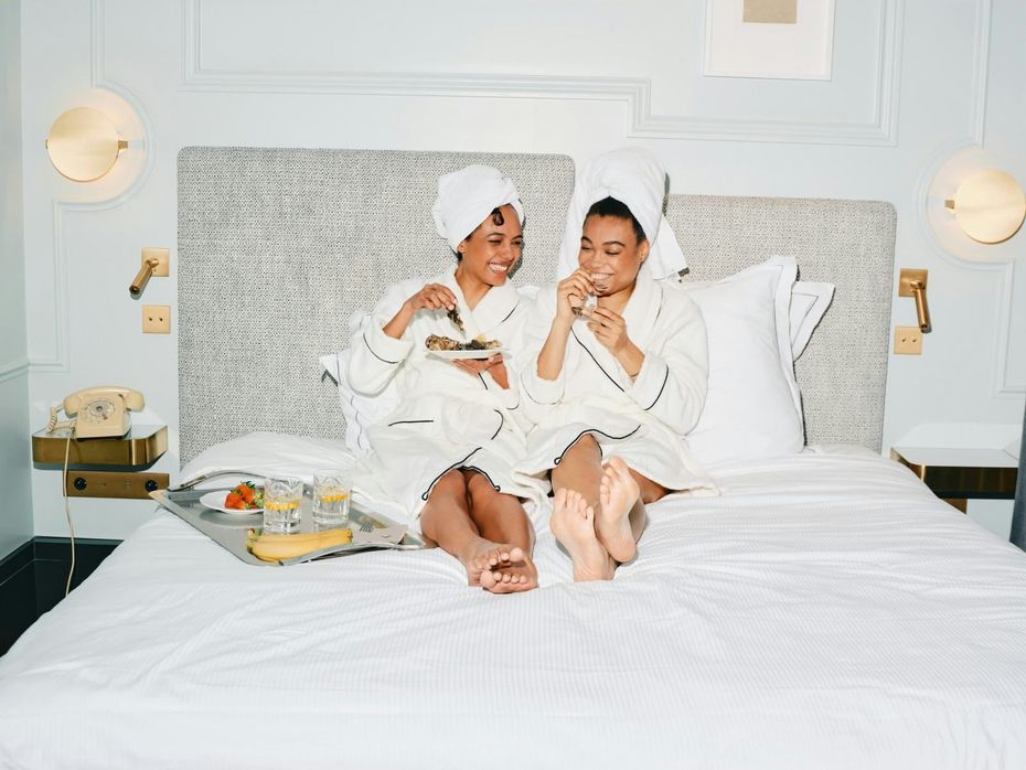 Two people in white robes sit on a bed, smiling and eating breakfast together.