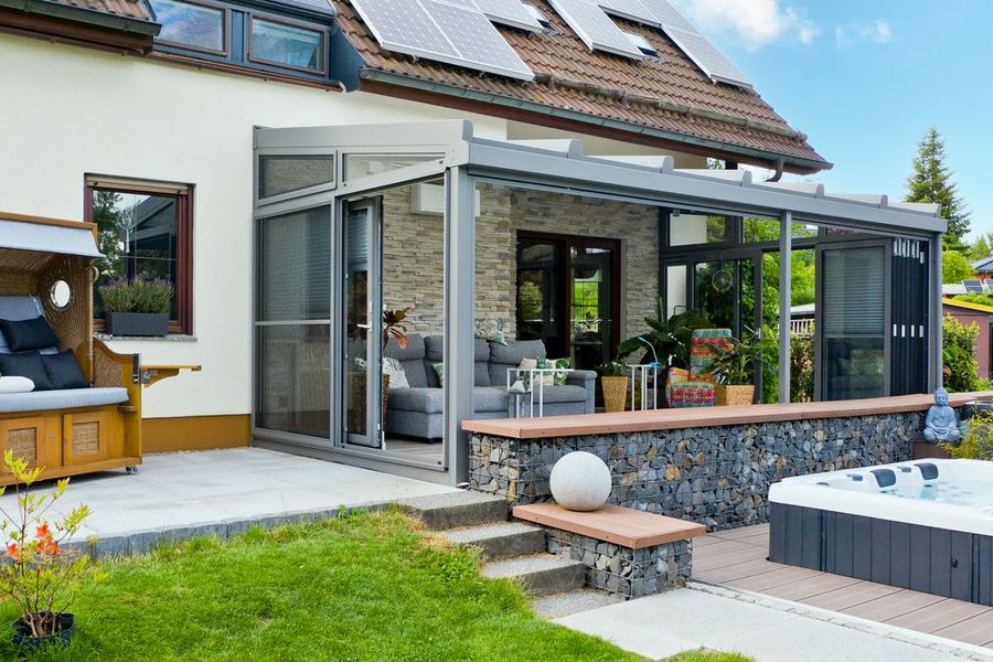 Moderner Wintergarten mit Steinmauer, Terrasse, Whirlpool und Sitzgelegenheiten, neben einem Haus mit Solarmodulen.