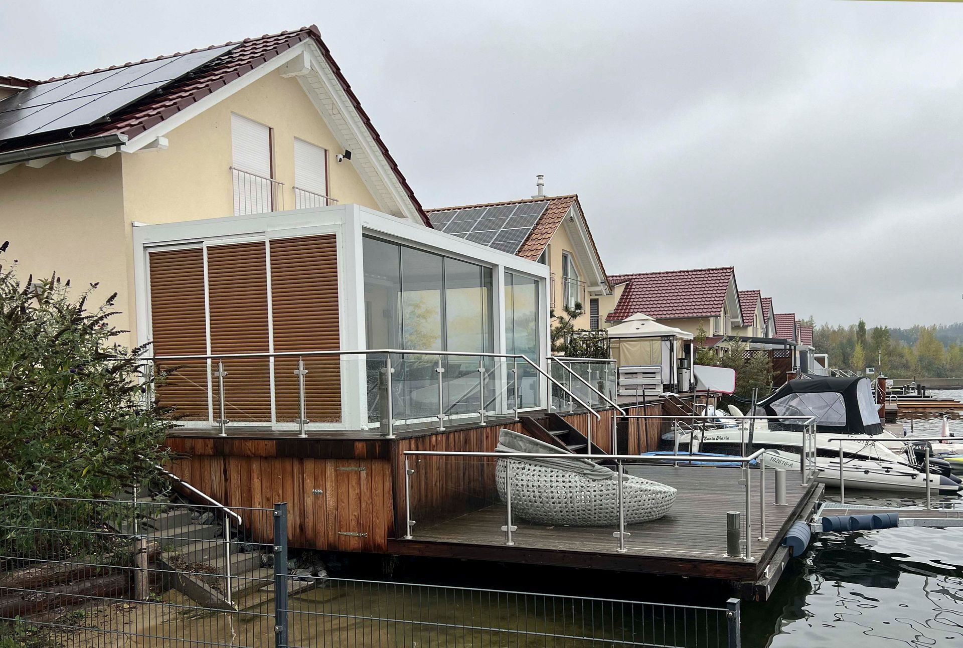 Schwimmendes Deck mit Glasumrandung und umliegenden Häusern auf einem See unter bewölktem Himmel.
