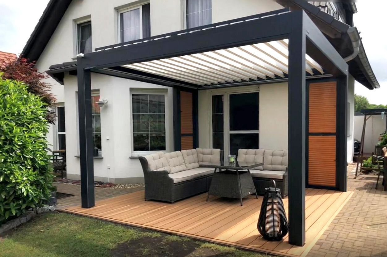 Eine Pergola mit dunklem Rahmen über einer Terrasse mit einer beigen Sofagarnitur. Holzterrasse und Fensterläden, weißes Haus.