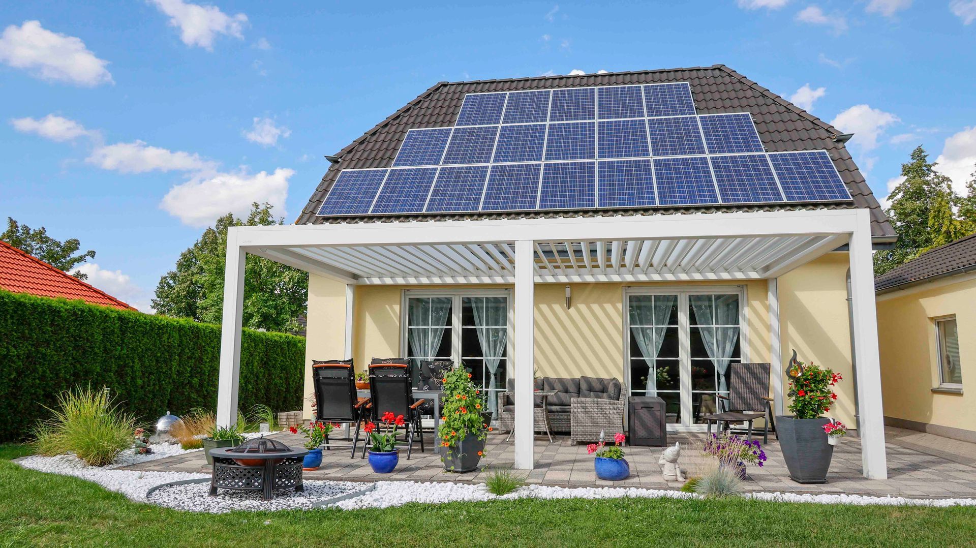 Haus mit Solarmodulen, Pergola und Sitzgelegenheiten auf der Terrasse unter blauem Himmel.