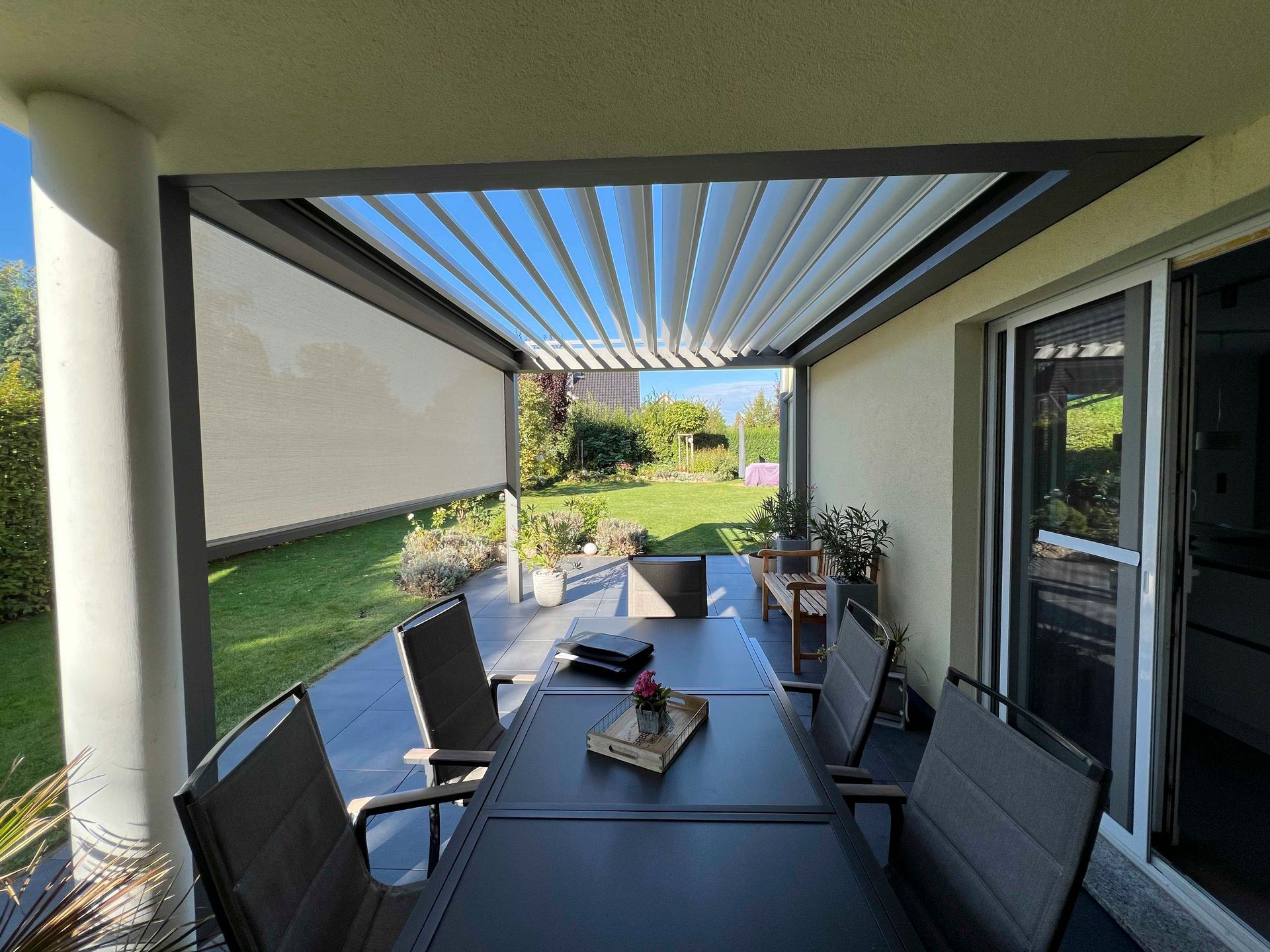 Terrasse mit Esstisch unter einem Lamellendach und Blick auf einen Garten.