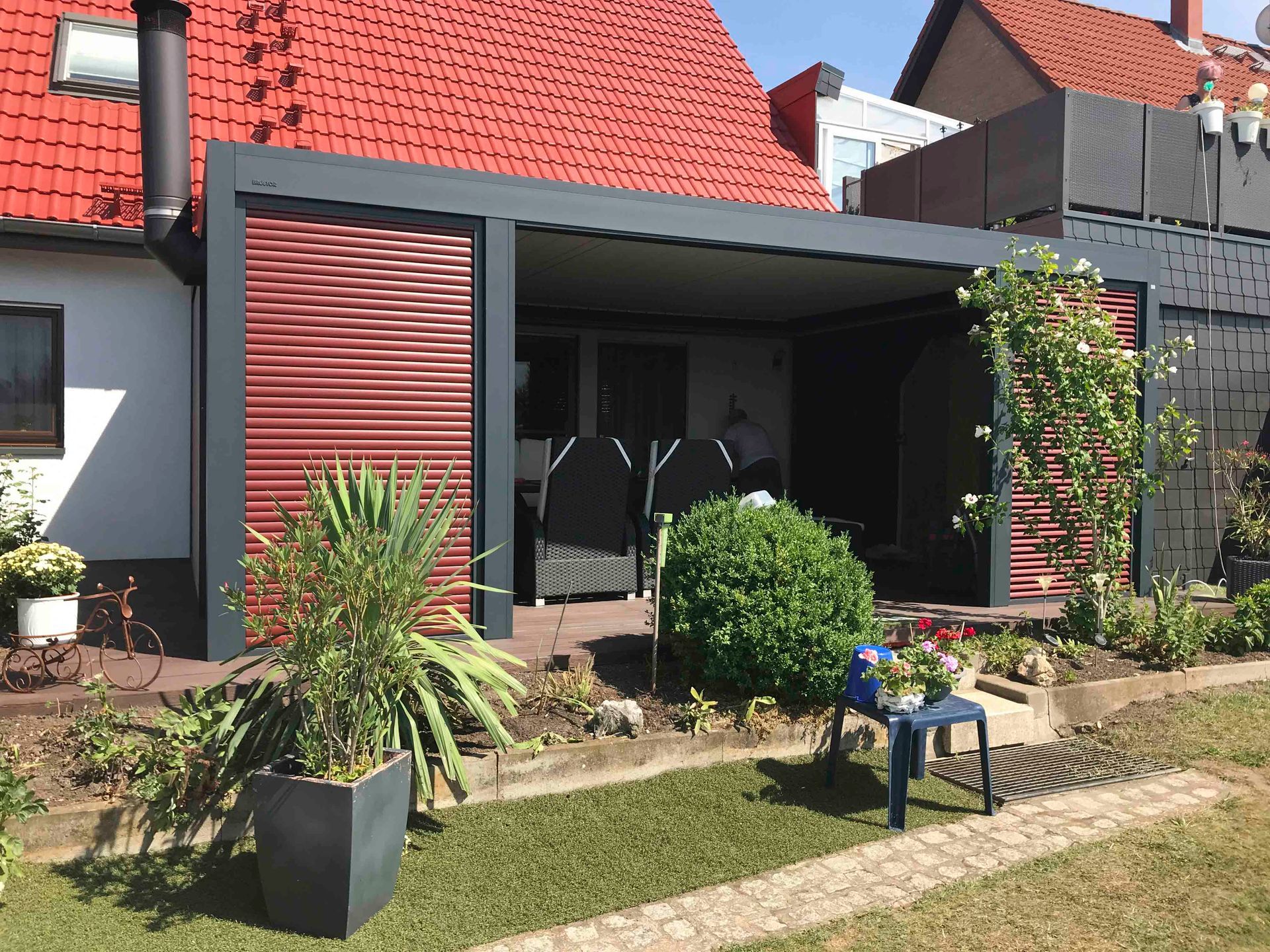 An einem Haus befestigte graue Pergola mit roten Lamellenseitenteilen.