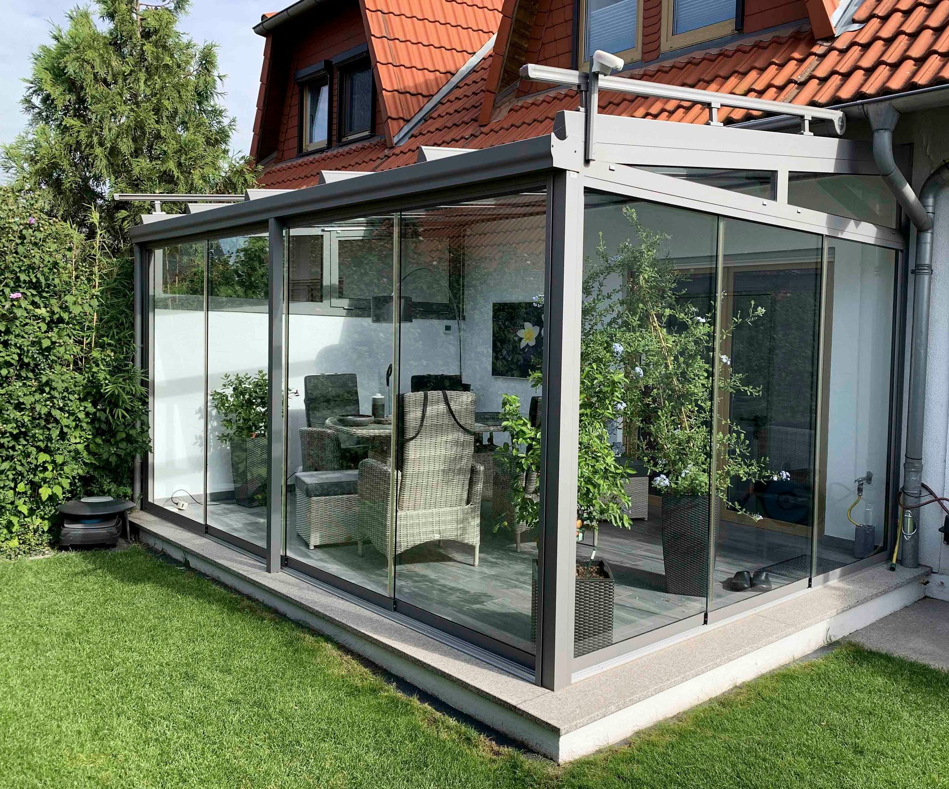 Angebauter Wintergarten mit Glaswänden an einem Haus, gefüllt mit Pflanzen und Möbeln, auf einer Rasenfläche.