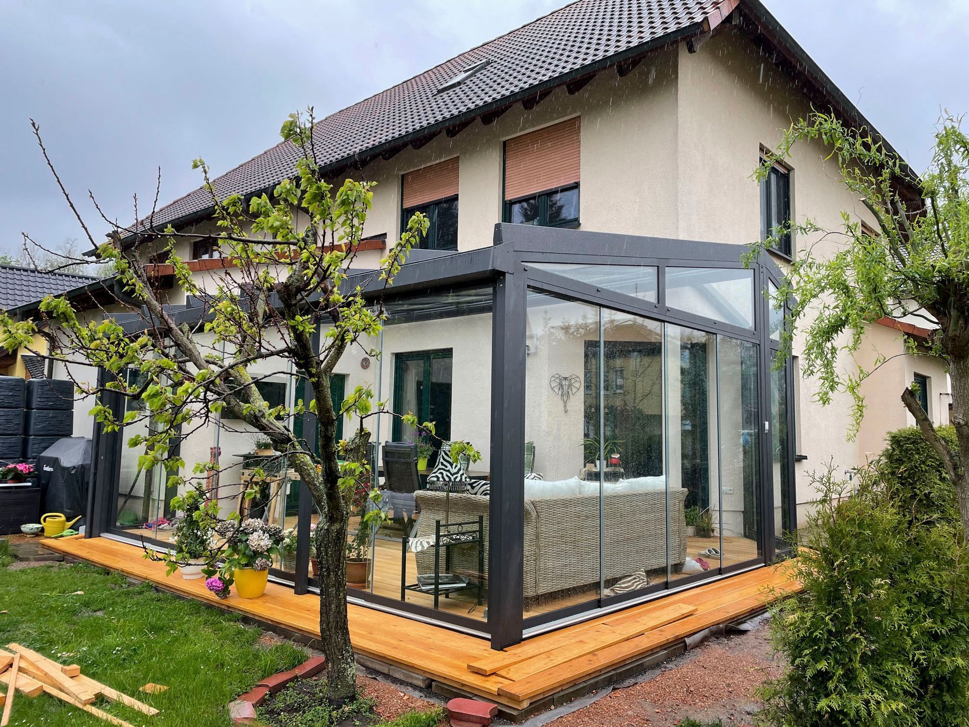 Geschlossener, verglaster Terrassenraum an einem hellen Haus mit Holzterrasse und grünem Rasen.
