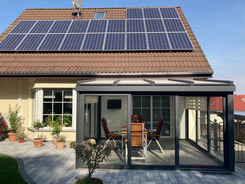 Haus mit Solarmodulen auf dem Dach, einem verglasten Wintergarten und einer gepflasterten Terrasse.