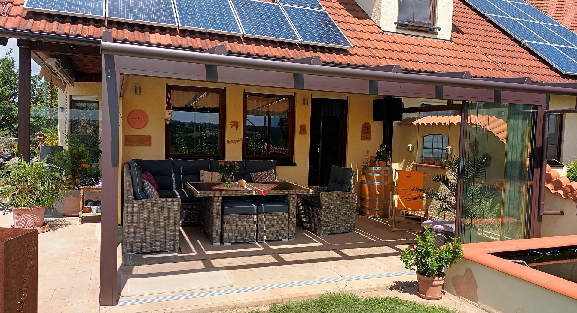 Außenterrasse mit Sitzgelegenheiten unter einer Pergola neben einem Haus mit Solarmodulen.