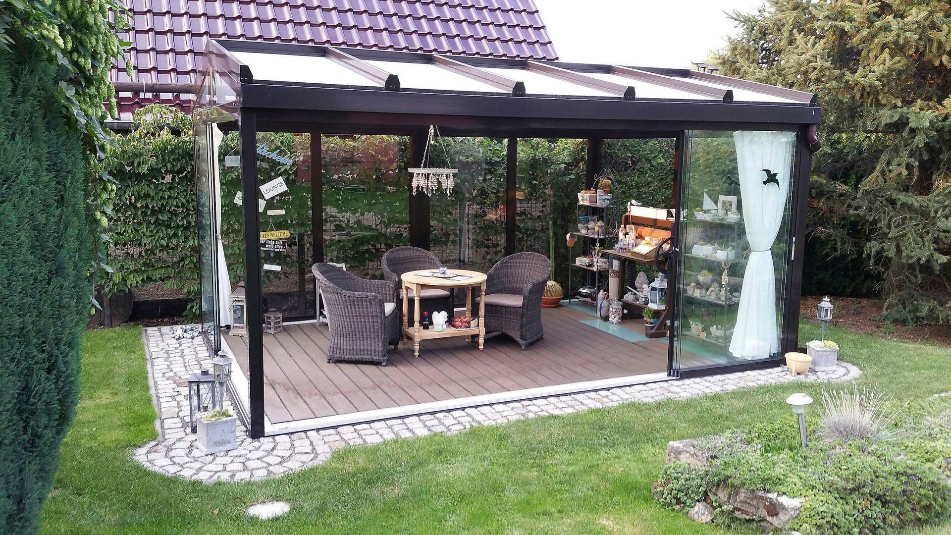 Verglaste Terrasse mit dunklem Rahmen, Korbmöbeln und Dekor. Auf einem Holzdeck, umgeben von Grün.