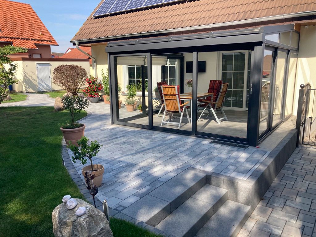 Wintergarten mit grauer Terrasse, Stufen und Essgruppe; Solarmodule auf dem Dach.