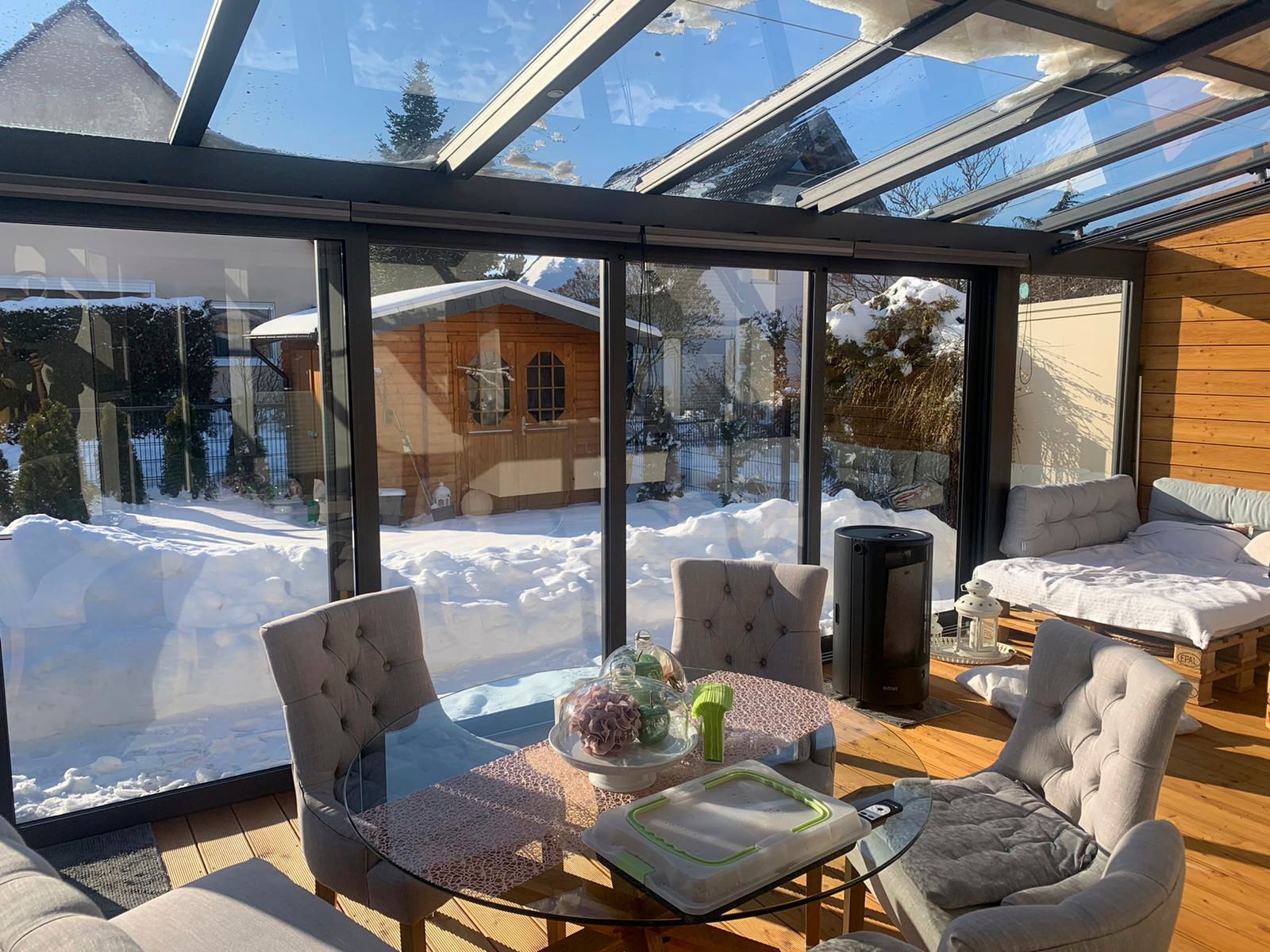 Wintergarten mit Holzschuppen und Blick auf den schneebedeckten Garten. Graue Stühle umgeben einen Glastisch.
