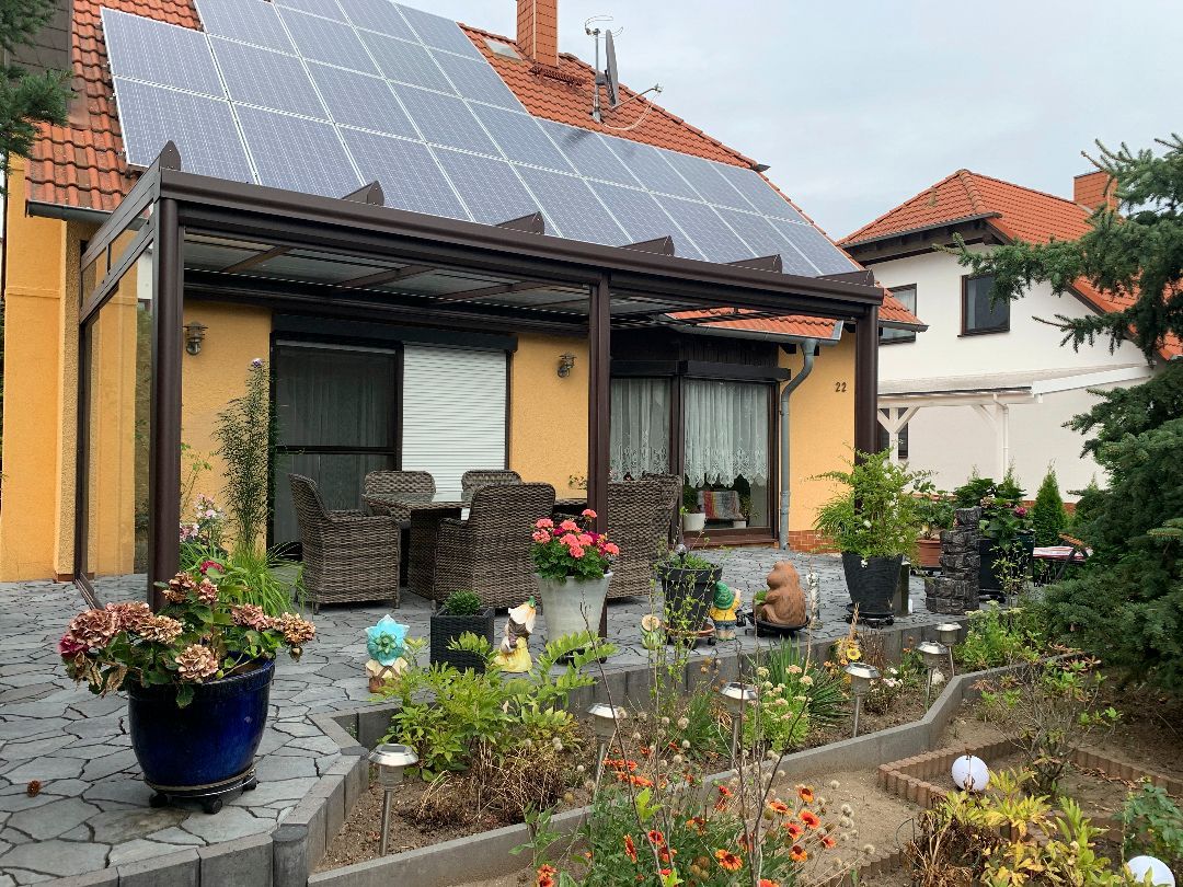 Gelbes Haus mit Solarmodulen, einer mit einer Pergola überdachten Terrasse und einem Blumengarten.