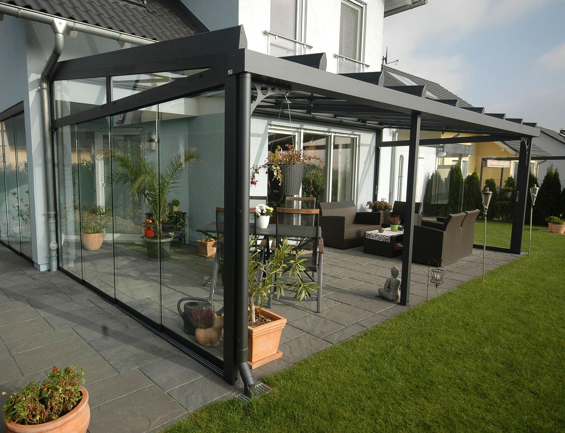 Verglaste Terrasse mit schwarzem Rahmen, Möbeln, Pflanzen, auf einer grauen Terrasse, neben einem grünen Rasen und einem Haus.