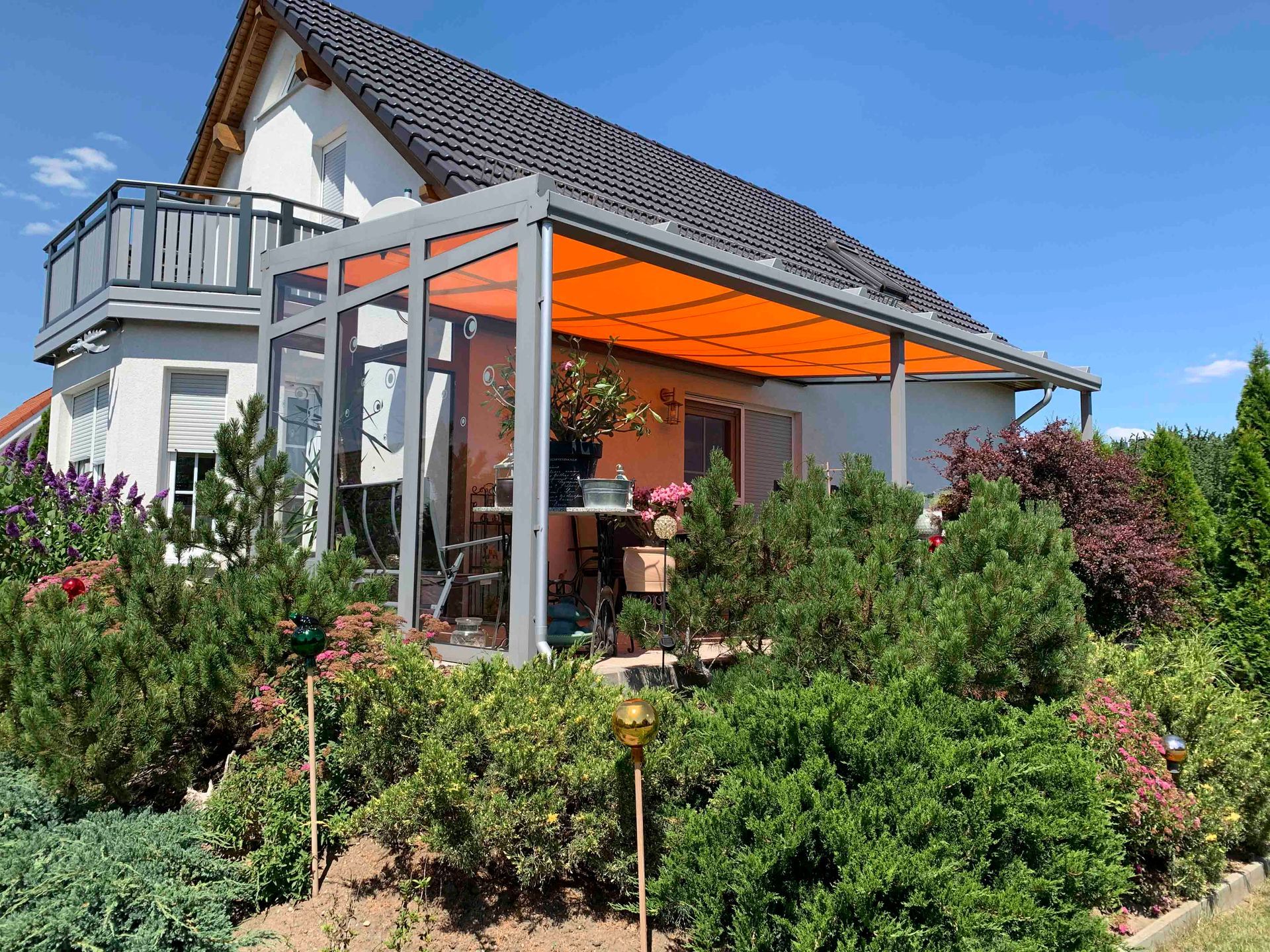 Ein weißes Haus mit einer orangefarbenen Markise mit grauem Rahmen über einer von grünen Büschen umgebenen Terrasse.