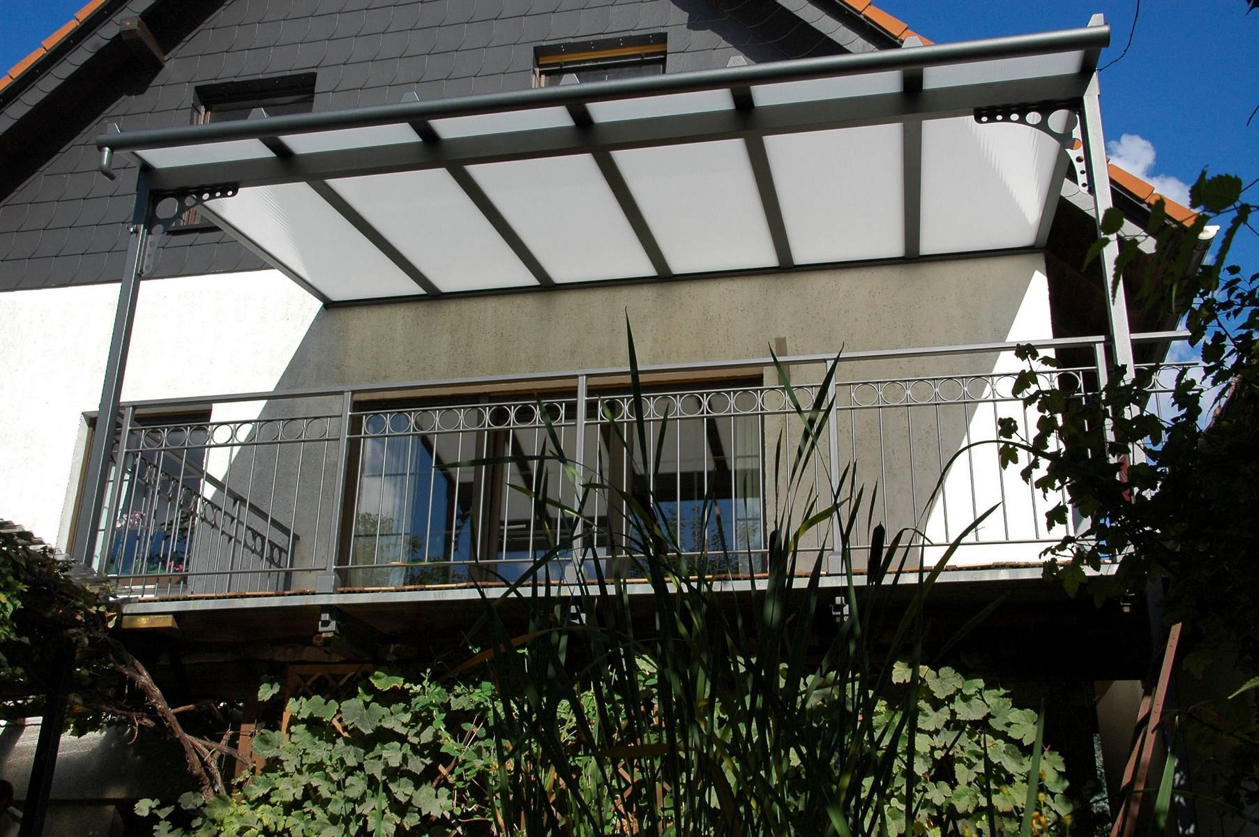 Balkon mit Metallrahmen, lichtdurchlässigem Dach und Geländer, von einem Garten aus an einem sonnigen Tag gesehen.
