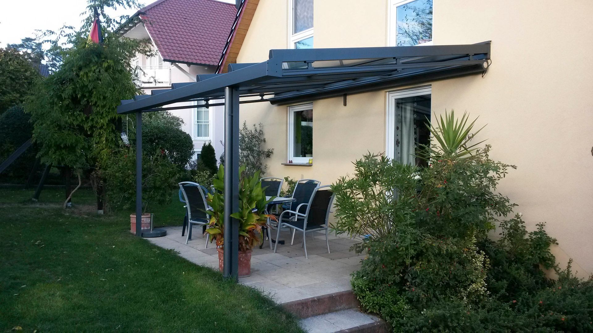 Terrasse mit Pergola mit dunklem Dach, Tisch, Stühlen und üppigem Grün neben einem gelben Haus.