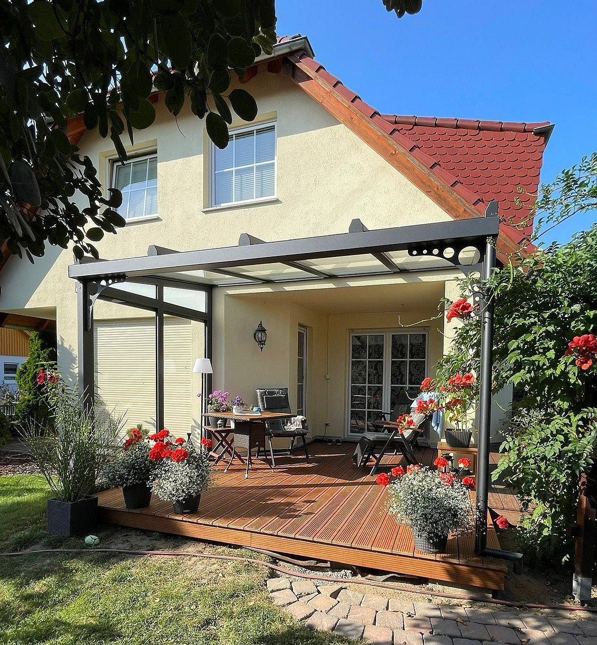 Holzterrasse mit Pergola an einem beigen Haus mit rotem Dach. Rote Rosen rahmen die Terrassenmöbel ein.